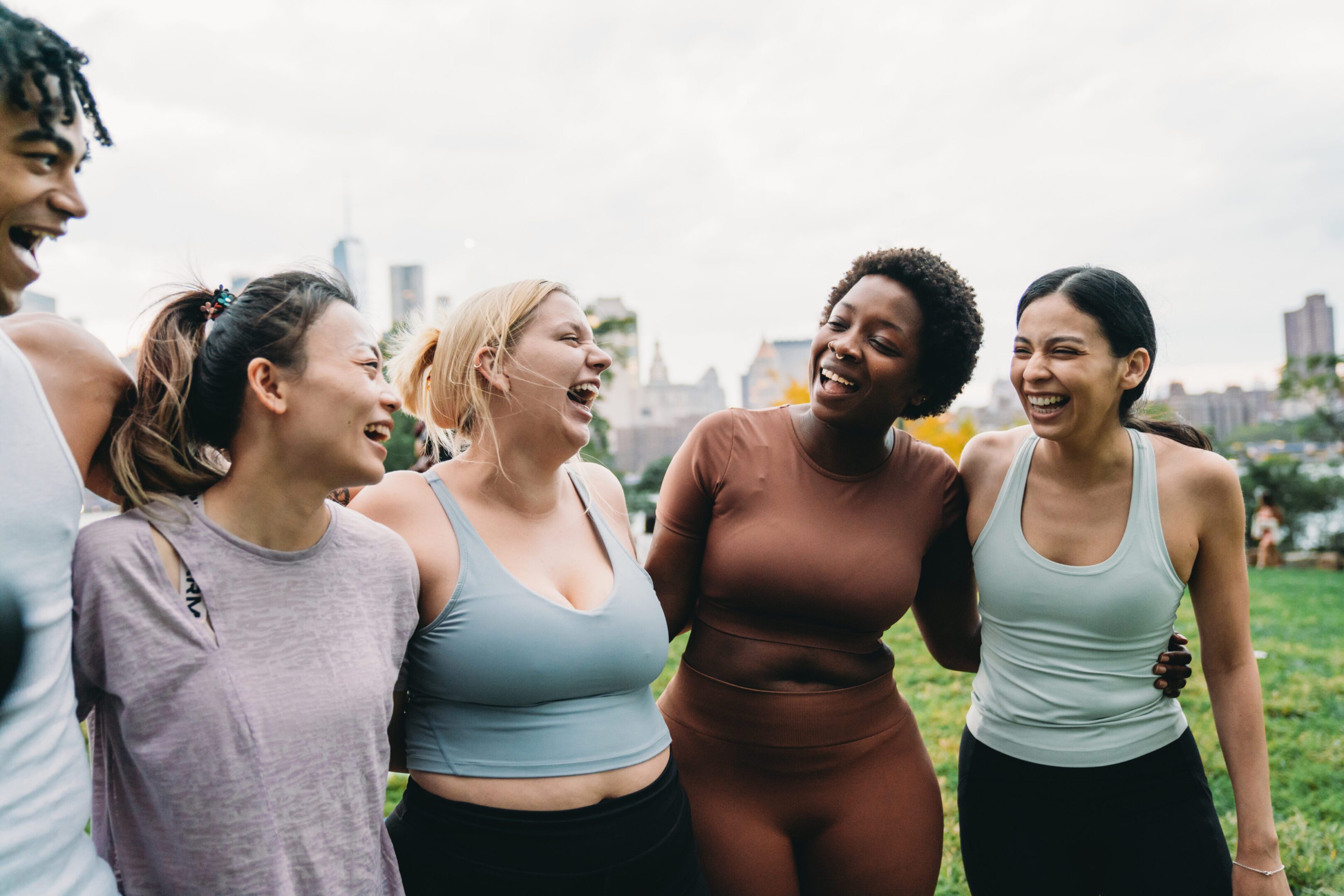 Four diverse women laughing together outdoors in athletic wear.